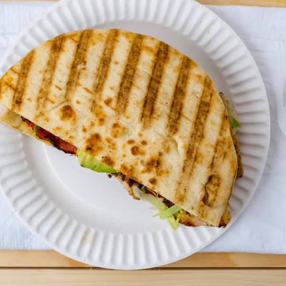 Grilled quesadilla with meat, lettuce, and avocado on a white paper plate.