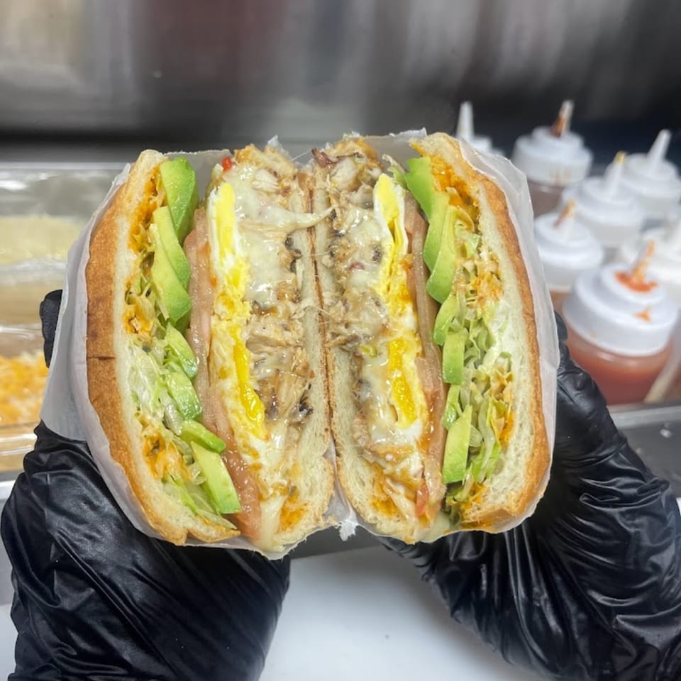 Hands in black gloves hold a sliced Bodega Bros deli sandwich filled with meat, cheese, avocado, and egg.