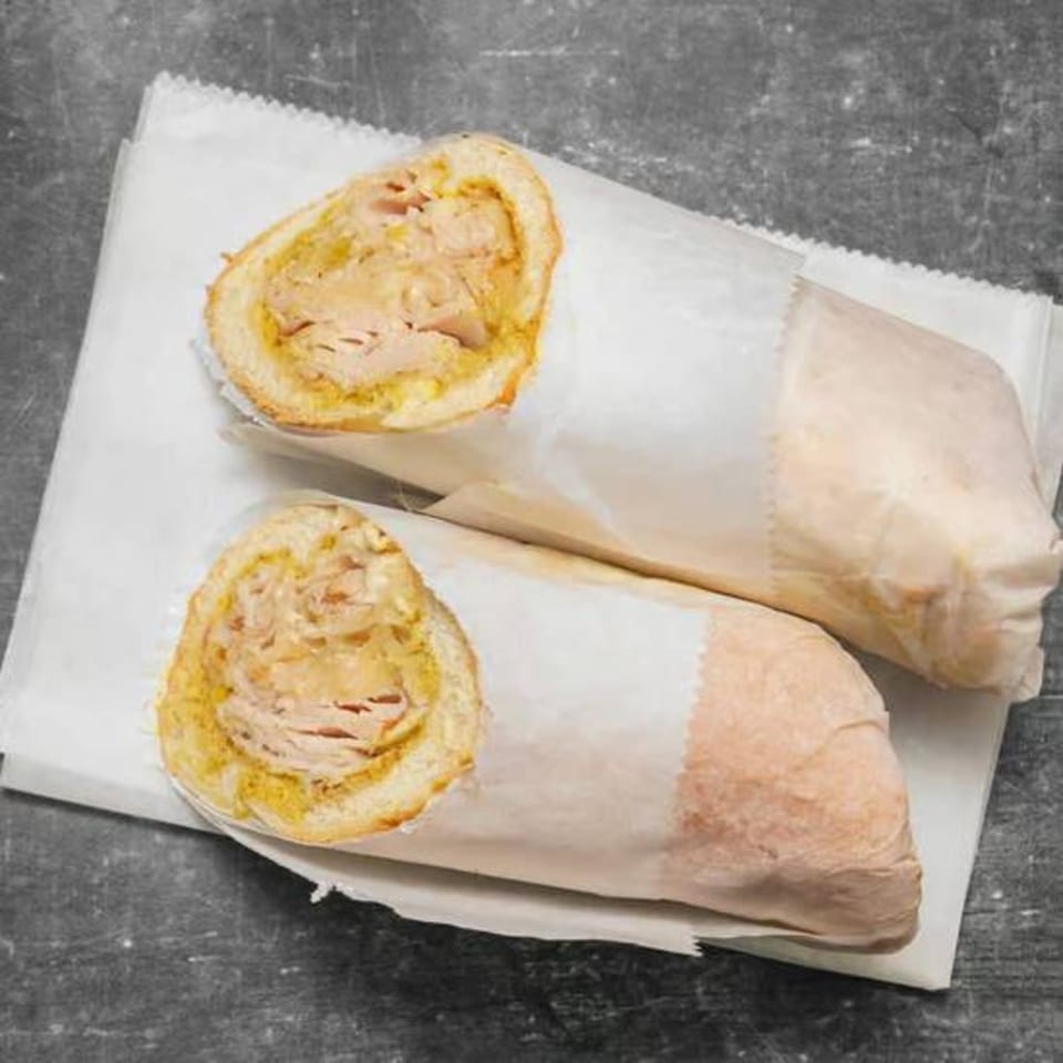 Two Bodega Bros deli sandwiches, filled with meat and mustard, wrapped in white paper on a dark surface.