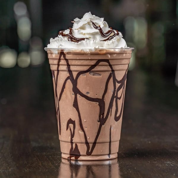 Chocolate milkshake with whipped cream and chocolate drizzle in a plastic cup on a dark table.