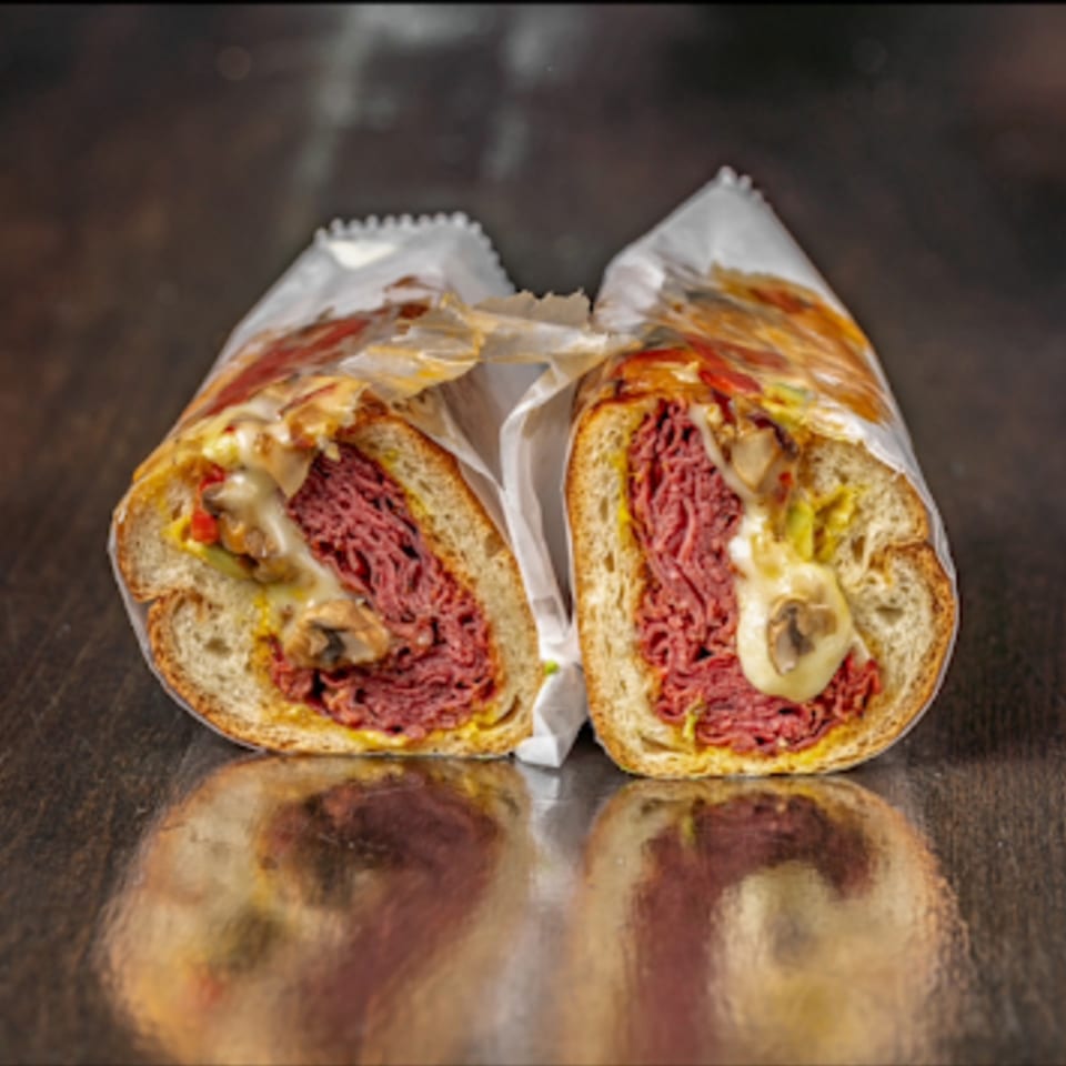 A Bodega Bros deli sandwich, cut in half, revealing pastrami, melted cheese, and peppers.