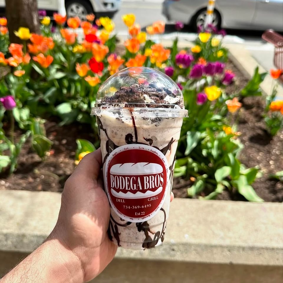 Hand holding a Bodega Bros milkshake with chocolate drizzle and cookie crumbles, against a backdrop of colorful tulips.